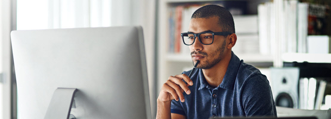 Man with close-cropped hair, beard and moustache wearing black glasses and medium blue short-sleeved polo shirt looking at computer monitor.