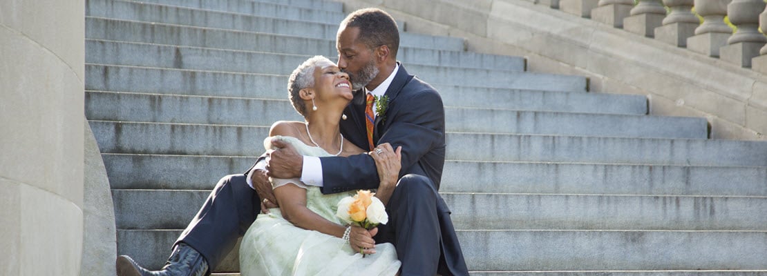 Older Black man dressed in a suit and cowboy boots and Black woman dressed in a pale green evening gown sitting on a granite staircase and hugging.