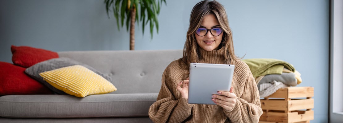 Woman with dark blonde long hair, wearing glasses and a tan ribbed sweater sitting in front of a light gray couch using a white tablet computer.