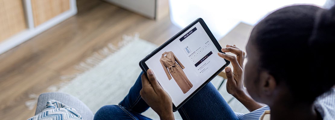 Image over the left shoulder of a Black woman looking at a tablet computer showing an image of long, tan coat for sale on a website