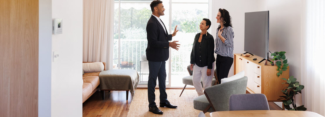 Tall man in the small, modern living room of an apartment discussing it with two women in front of a television on a credenza.