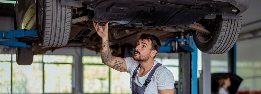 Young, bearded mechanic in light blue t-shirt and blue overalls touching underside of car with his right hand; the car raised up on a garage lift