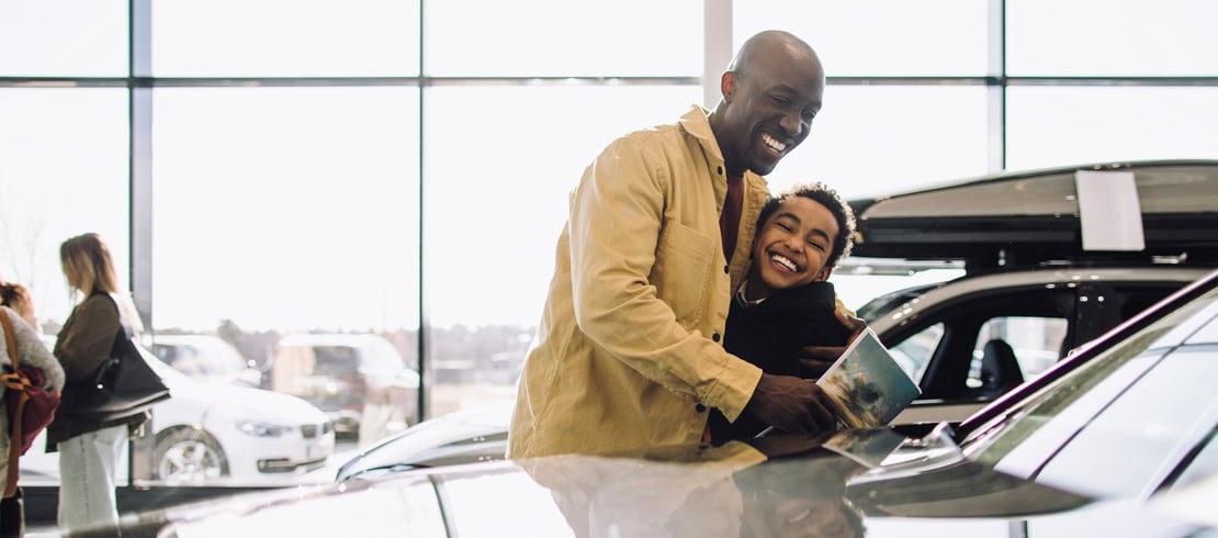 man smiling looking at a new car on a dealership show room while hugging a child