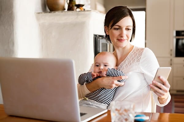 mother at home with baby working on laptop, holding smartphone