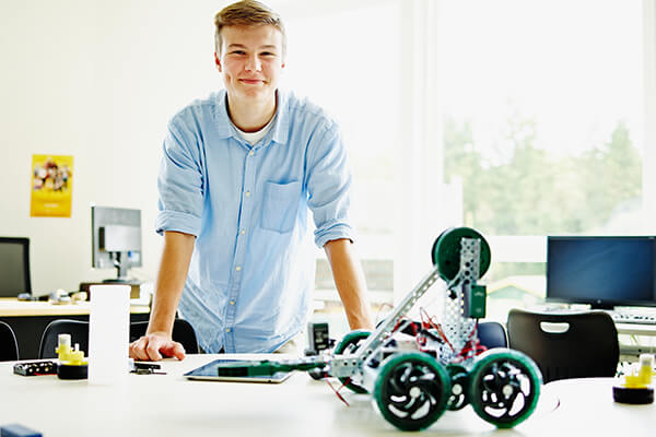 smiling male student leaning on table with robotics project and digital tablet in high school classroom