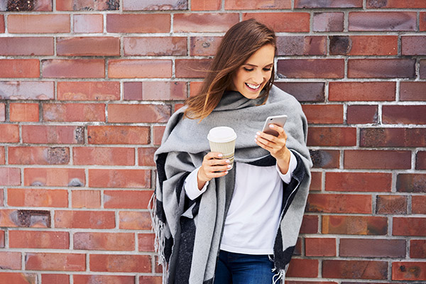 happy woman with coffee to go looking at cell phone in front of brick wall